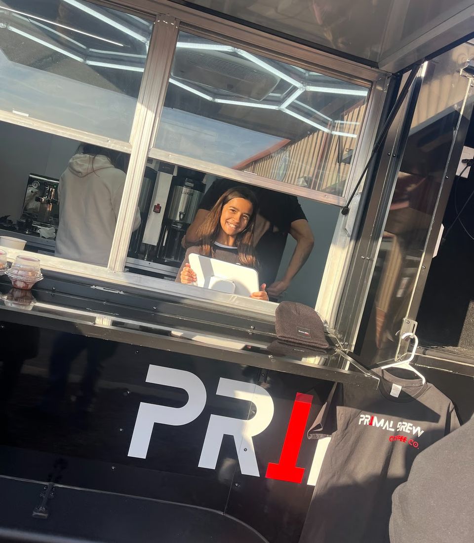 Barista serving customers from the window of a Primal Brew coffee trailer